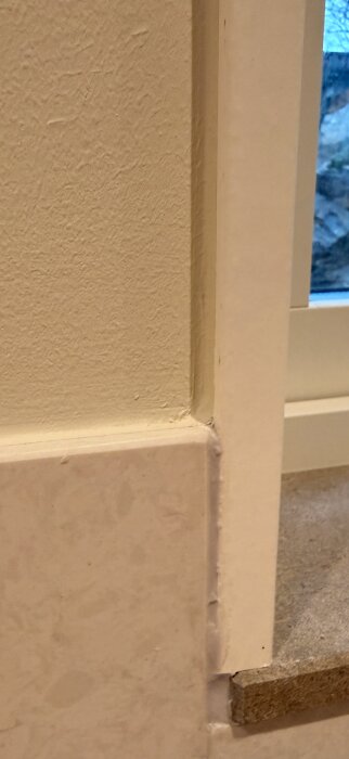 Nära bild på ett hörn där en vägg, fönsterram och list möts, med ojämn och slarvig målning eller tillpassning.