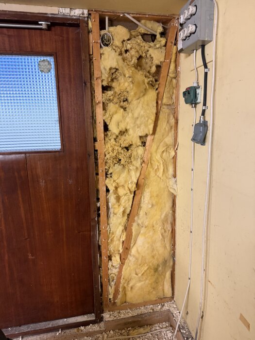 Öppen vägg med isolering som delvis har rivits, visar träreglar och elkablar. Väggens innandöme i ett gammalt garage från 70-talet.