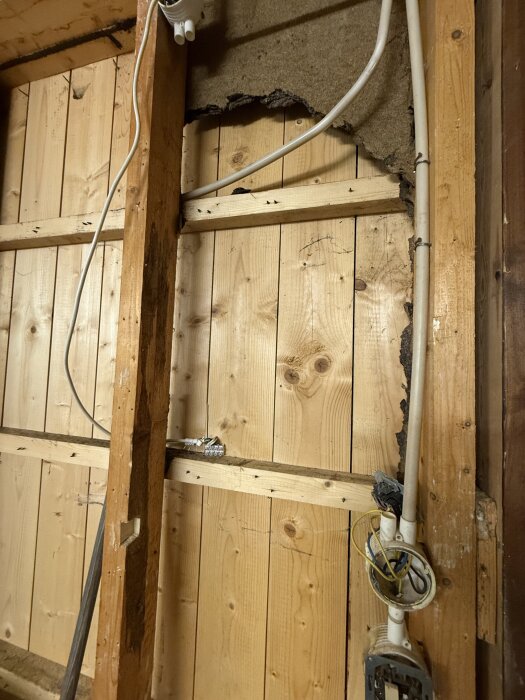 Vägg av träreglar och isolering med skador, exponerade kablar och tecken på fuktproblem i ett garage från 70-talet.