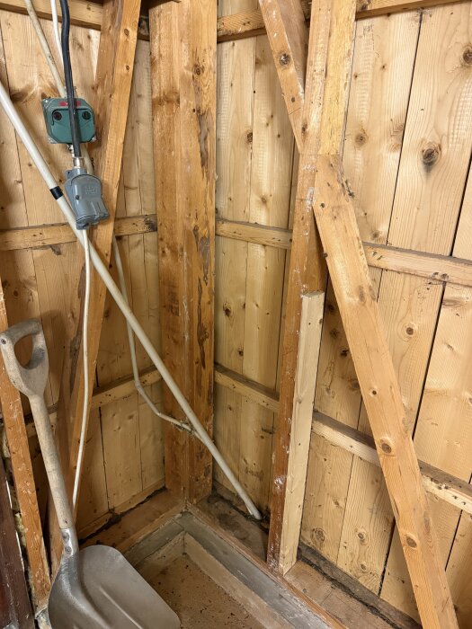 Innersta delen av ett garage med träreglar, eluttag och en spade. Synliga fuktskador och missfärgningar på trästommarna.