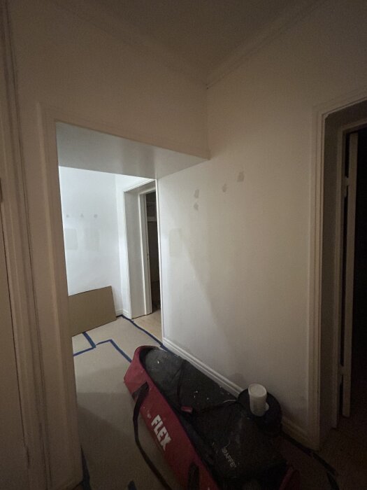 En smal hall med vita väggar, markerade golvlinjer och en röd verktygsväska på golvet. Stolar och rengöringsprodukter står längs väggen.