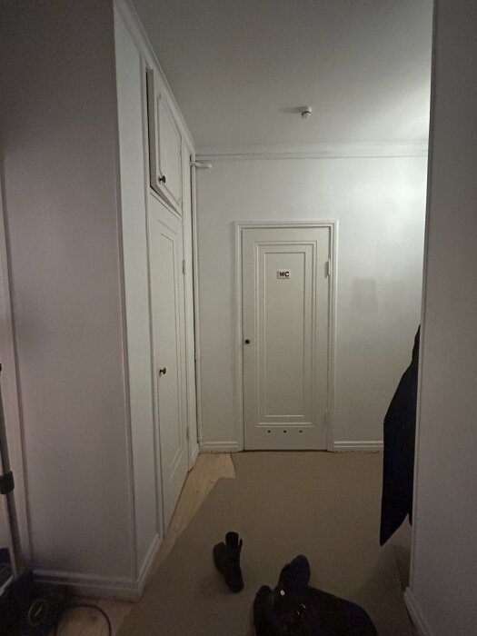 Hall med vitmålade väggar och tak, en dörr med "WC"-skylt, beige golv och en stövel på golvet.