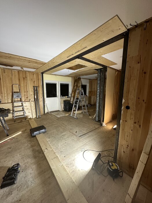 Renoveringsområde med synlig träram och stålbalk, stegar och bygglampor. T-balken är en del av ett byggprojekt, med fokus på eldragning.