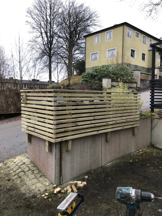 Trästaket byggt ovanpå en betongmur vid en gata, med en gul byggnad och höga träd i bakgrunden. Verktyg och tegelstenar syns på marken.