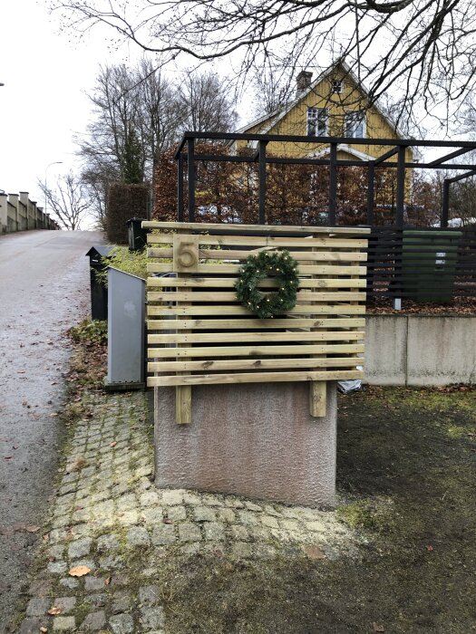 En nytillverkad träram med husnummer 5 och en grön krans, placerad framför en gul byggnad på en trottoar.