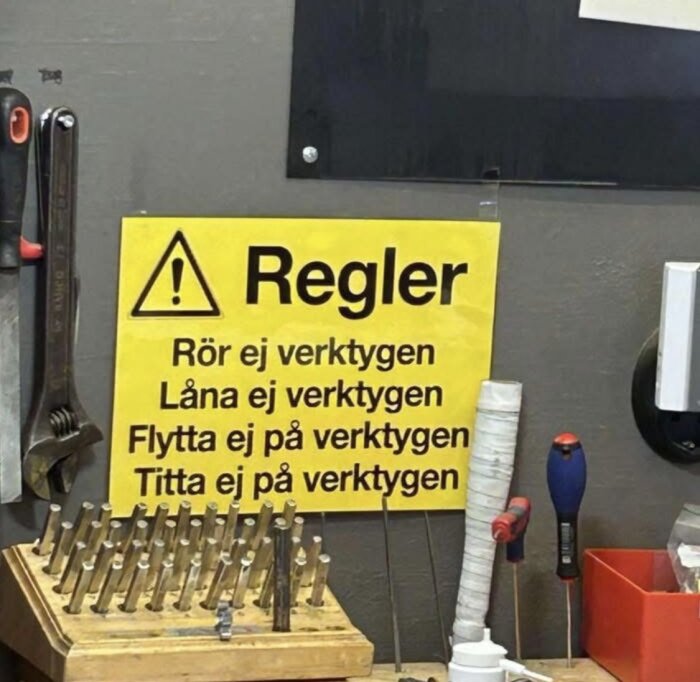 Verkstadsvägg med gult anslag om regler för hantering av verktyg; rör ej, låna ej, flytta ej, titta ej. Verktyg som tång och skruvmejslar syns.