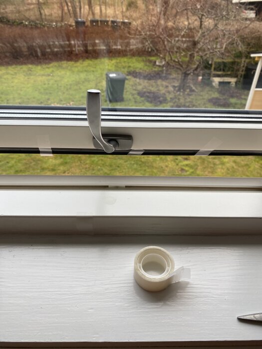 Elite fönster med aluminiumram i vädringsläge, handtag synligt, med lös tejp på karmen, samt rulle tejp och en sax på fönsterbrädan.