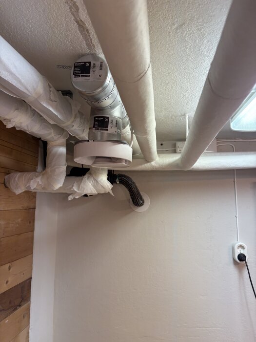 Vitmålad fläkt och rörinstallation i ett rum med träpanel och vit vägg; fläkten har monterats på väggen men behöver ytterligare arbete.