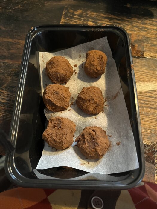 Sex hemmagjorda chokladtryffelbollar på bakplåtspapper i en plastlåda, täckta med kakao.