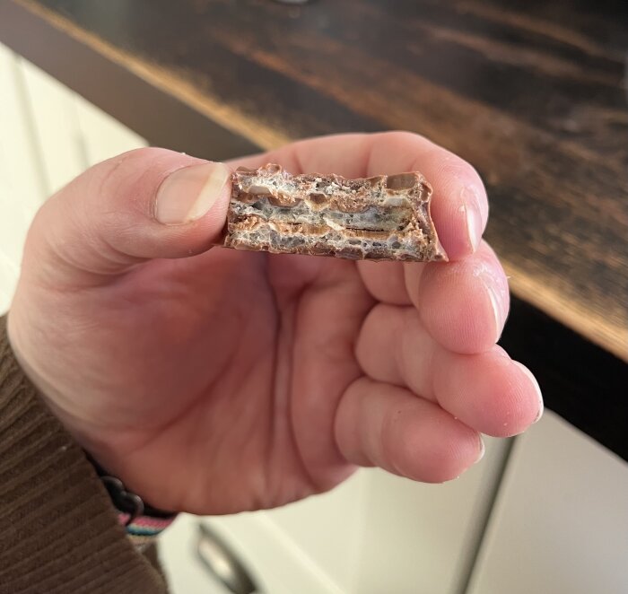 En hand håller en bit hemgjord kexchoklad med lager av choklad och jordnötssmör mellan smörgåsrån, delvis bitet och fotat i köksmiljö.