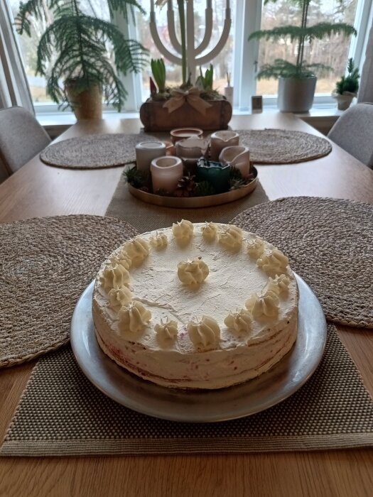 Gräddtårta dekorerad med gräddtoppar på ett fat, placerad på ett matbord med rottingunderlägg och ljusdekorationer i bakgrunden.