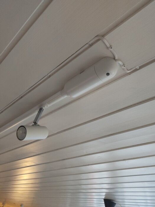 Vit ljusskena med spotlampa på vit träpaneltak, med en avlång enhet i slutet som kan innehålla en transformator.