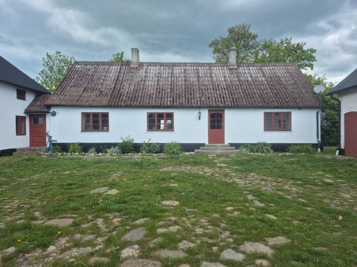 Vit fasad av lantligt hus med rött tak, röd dörr och fönster. Ogräsfri innergård med gräsmatta och stenläggning i förgrunden. Molnig himmel.