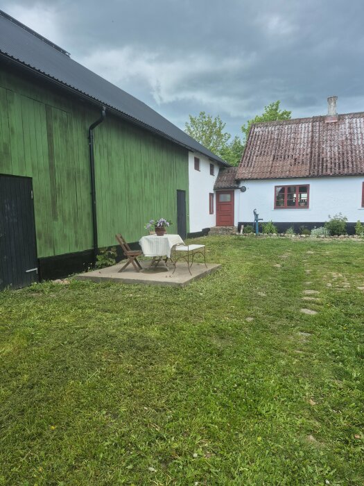 En rensad innergård med grönt träskjul, vitkalkat hus och stenlagd uteplats med ett bord och stolar, omgiven av nyslagen gräsmatta.