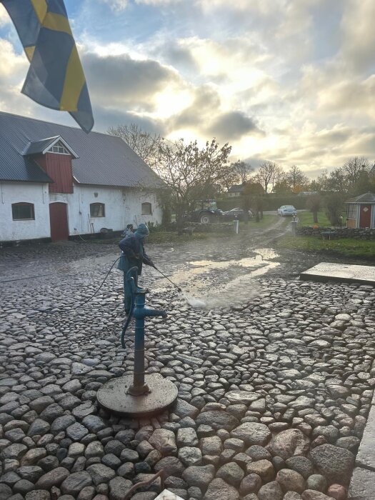 En stenlagd innergård rensad på ogräs, med en vattenpump i förgrunden. Svensk flagga och en gårdsbyggnad syns i bakgrunden.