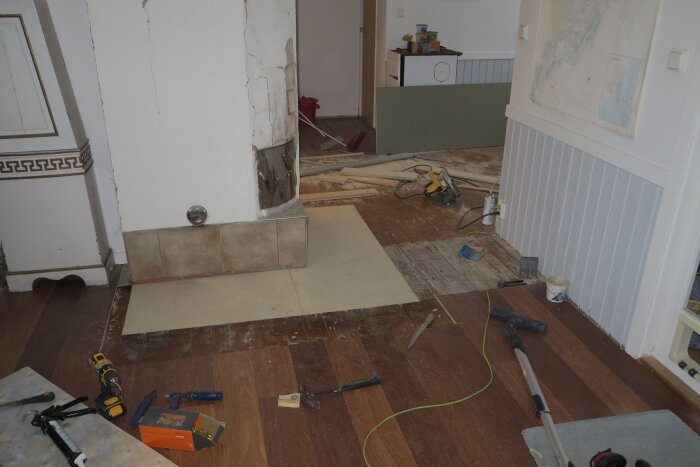 Renovering av golv med utrustning som skruvdragare, tapetklister och såg utspridda. Braskamin syns i bakgrunden. Golvplankor och verktyg på golvet.
