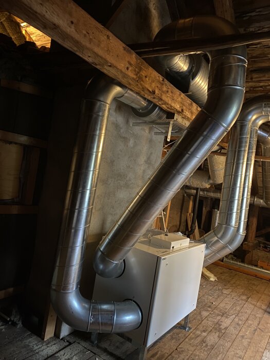Ventilationsrör i metall installerade på en vind, anslutna till en rektangulär enhet placerad på golvet av träplankor.