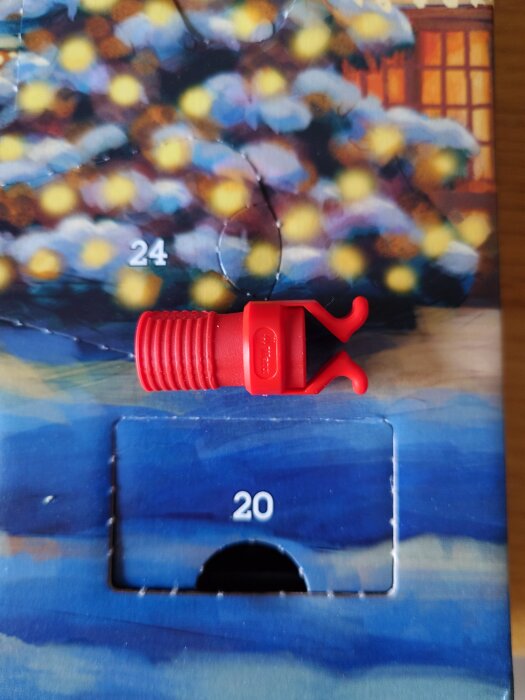 Röd plastkomponent ligger ovanpå en adventskalender med nummer 20 öppnat, bakgrunden är en suddig vinterbild med julgransmotiv och siffran 24.
