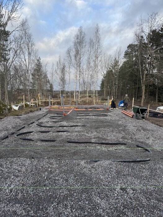 Grusbelagd byggplats med avloppsrör på plats i skogsmiljö, redo för nästa steg med el och bredbandsslang innan isolering.