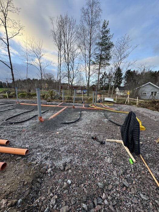 Byggarbetsplats med nylagda avloppsrör, grusad mark och omgivande träd. Redo för eldragning och bredbandsslang.
