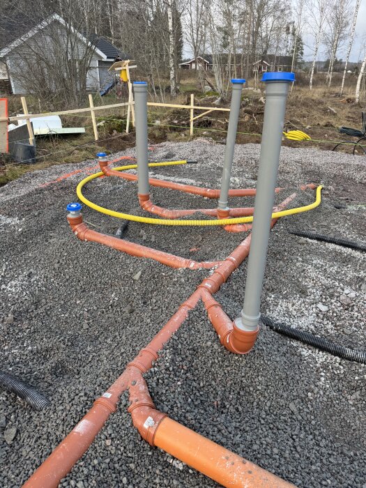 Avloppsrör installerade med korrekt lutning i ett byggprojekt, på grusbelagd mark omgiven av träd och en husfasad i bakgrunden.