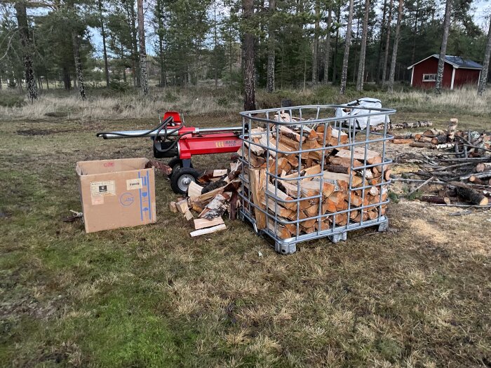 Rödklyv, vedkorg med grovkluven ved av gran, björk och tallar samt kartong på gräsmatta omgiven av skog.