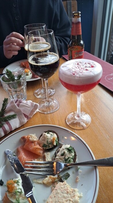 Tallrik med smårätter, glas med mörk dryck och rosa drink med skum står på träbord, tillsammans med flaska julmust och rödvit servett.