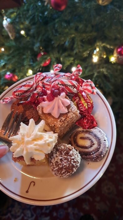 En tallrik med julgodis och bakverk framför en julgran, inklusive chokladbollar, rulltårta, små kakor och godis med inslag.