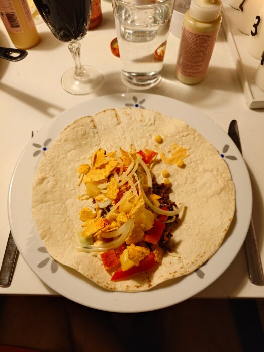 Taco med köttfärs, lök, paprika, majs och tortillachips på en tallrik bredvid ett glas vin och ett glas vatten.