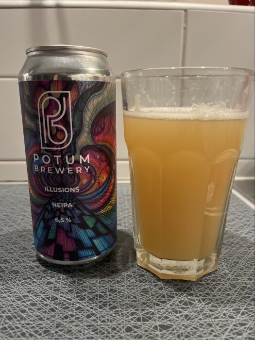 Burk och glas med Potum Brewery Illusions NEIPA, en grumlig öl med 6,5% alkoholhalt, på ett mönstrat bord.