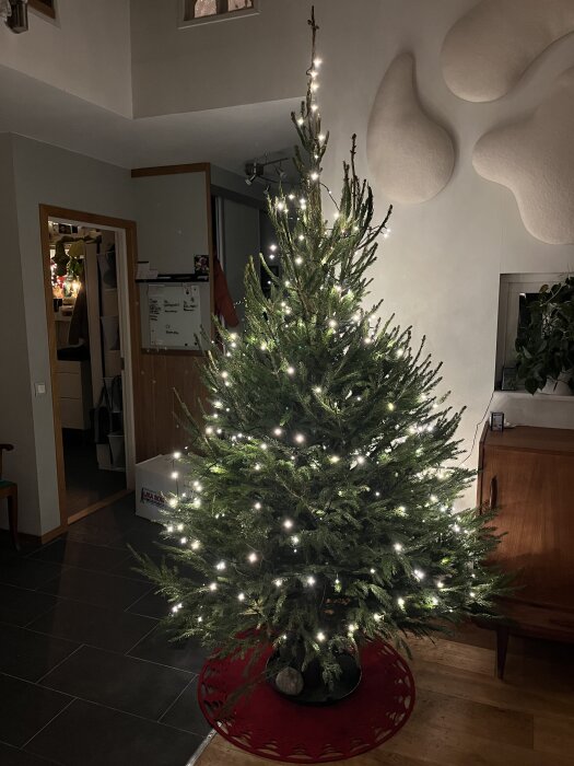 Julgran inomhus dekorerad med ljusslinga, men utan julkulor, stående på en röd matta i ett modernt vardagsrum.