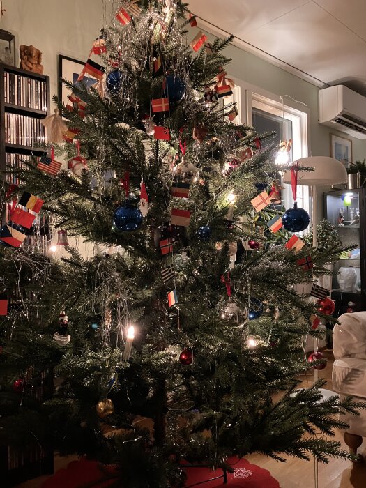 Julgran dekorerad med flaggor, blåa och röda kulor, silverglitter och ljus, placerad i ett vardagsrum med ljusinsläpp från fönster.