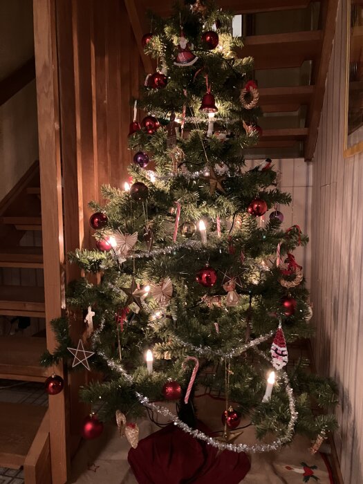 Julgran dekorerad med röda och silverfärgade kulor, halmdekorationer och ljus, placerad vid en trappa i ett hem.