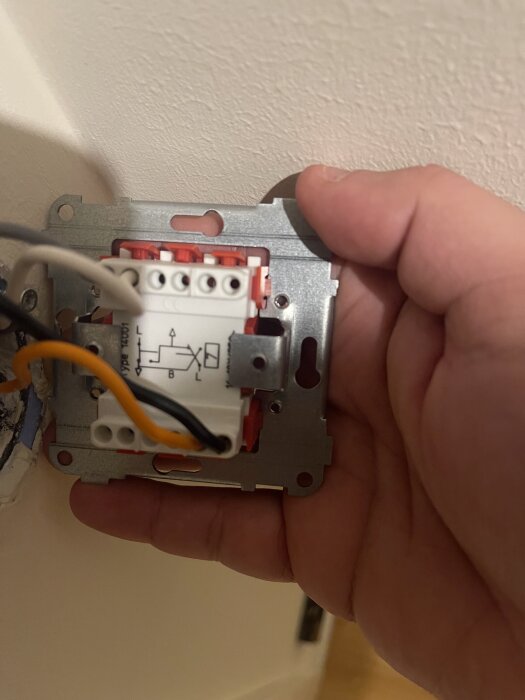 Hand håller en elektrisk strömbrytare monterad i väggen, med synliga kablar anslutna till enheten.