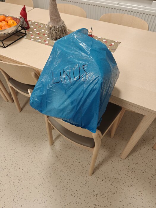 Blå plastpåse med texten "LINUS" placerad på en stol vid ett bord dekorerat med julduk och dekorationer.