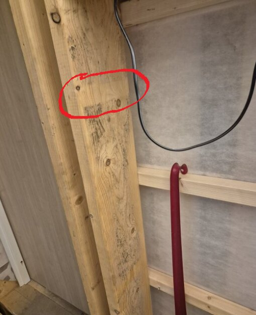 Träkonstruktion med två vertikala reglar, en röd krok och en svart kabel. Ett område är markerat med en röd cirkel på ena regeln.