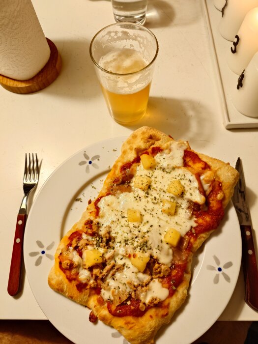 Pinsa med prosciutto, tonfisk, ananas och ost på tallrik, bredvid ett glas öl.