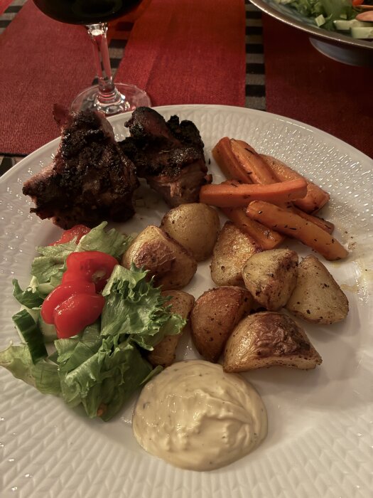 Tallrik med grillat kött, ugnsstekta potatisar, morötter, sallad och sås, serverat med rödvin.