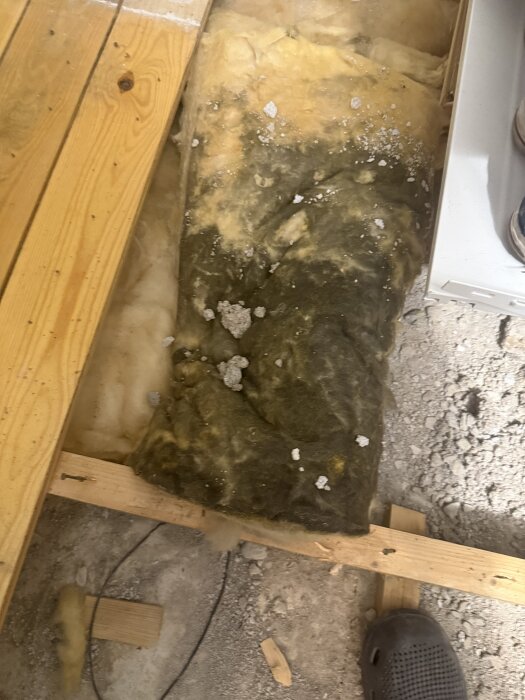 Isoleringsmaterial under golvplankor i gammal byggkonstruktion, delvis täckt av damm och skräp.