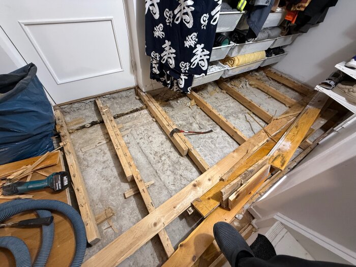 Bilden visar ett rum under renovering där gamla träbjälkar är synliga efter att golvet rivits upp. Verktyg och skräp finns runt omkring.