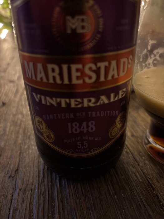 En flaska Mariestads Vinterale på ett träbord bredvid ett glas halvdrucken öl.