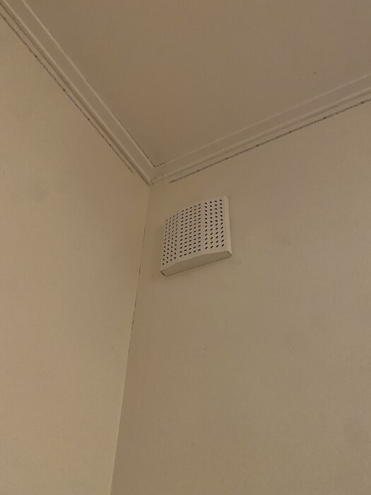 Väggmonterat frånluftsdon i ett hörn av ett rum med vanligt galler.