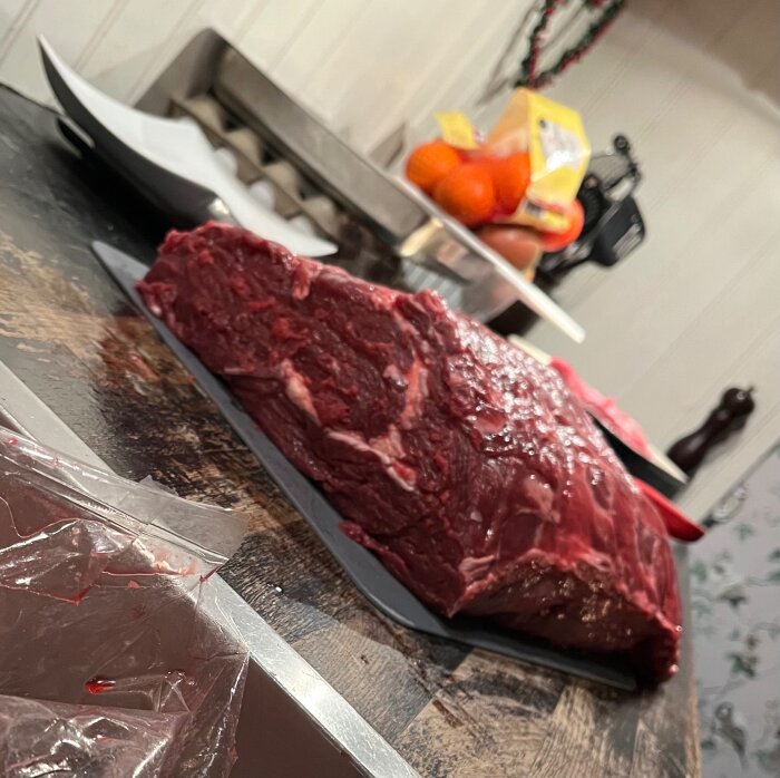 Stort stycke rått kött ligger på skärbräda på träbord med frukt och köksredskap i bakgrunden.