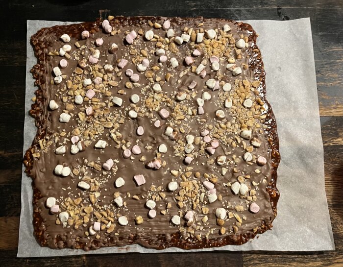 Hemmagjord chokladtäckta barer med mini-marshmallows och hackade nötter ovanpå, ligger på bakplåtspapper.