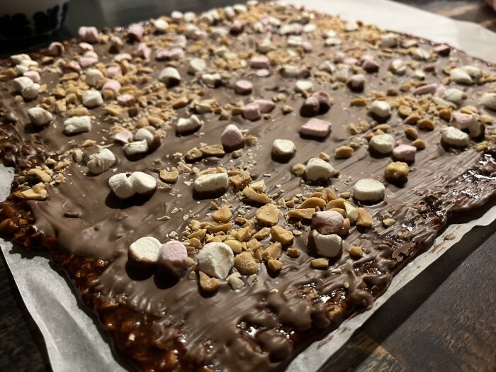 Hembakad chokladkaka toppad med små marshmallows och hackade nötter ligger på en bakplåtspappersklädd yta.