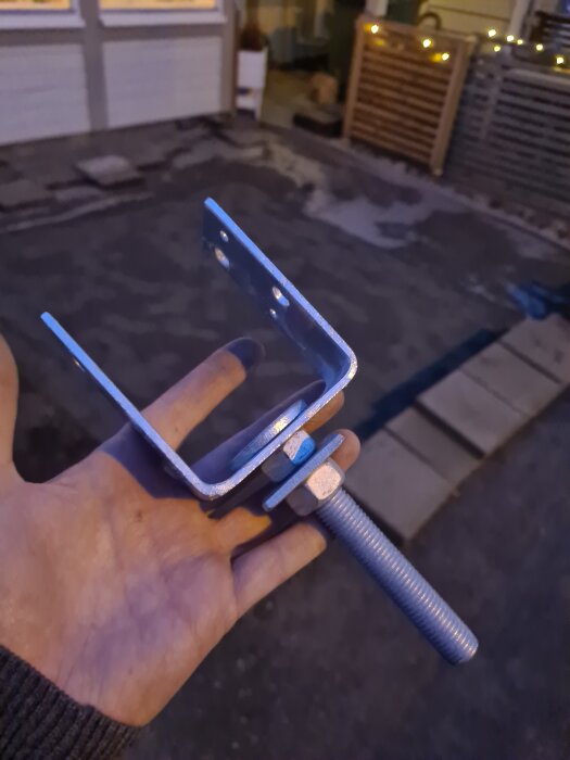Hand håller en metalldubb med en U-formad stolpsko och mutter, bakgrunden visar en trädgårdsinfart belagd med stenar och omgiven av en spaljé.