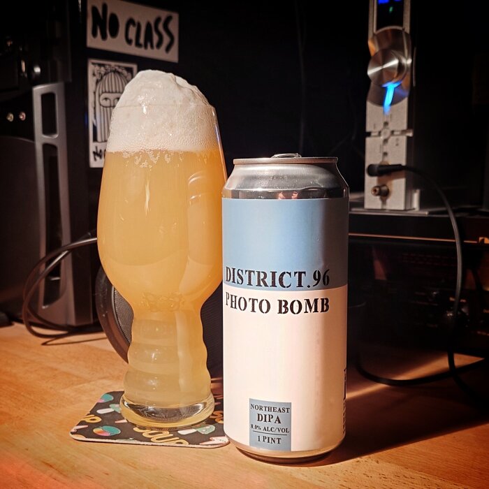 Ölglas med skummande öl bredvid en burk märkt "District 96 Photo Bomb" på ett bord.