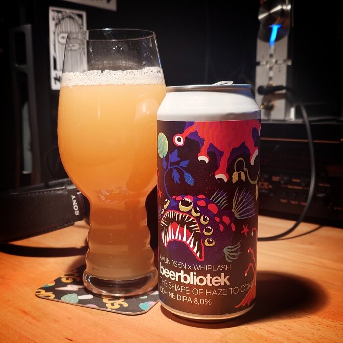 Ölglas med grumlig dryck bredvid en färgglad ölburk märkt "Beerbliotek The Shape of Haze to Come".