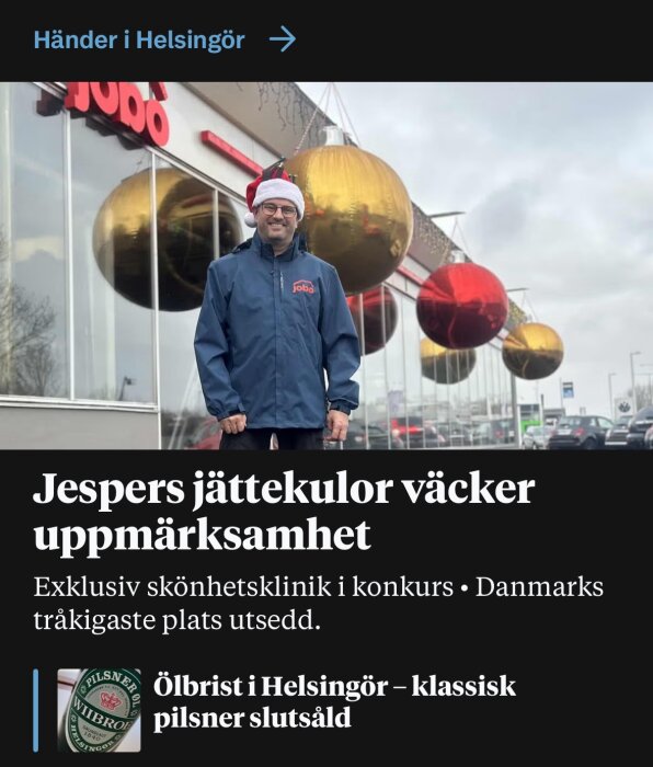 En man med tomteluva framför en byggnad dekorerad med stora julkulor och skylten "Jodo". Text om jättejuldekorationer och Helsingör.
