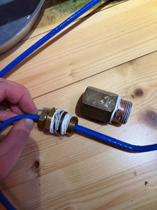 Blå och mässingsfärgade rördelar med gängor och tätningstejp på träbord, en hand håller i röret.